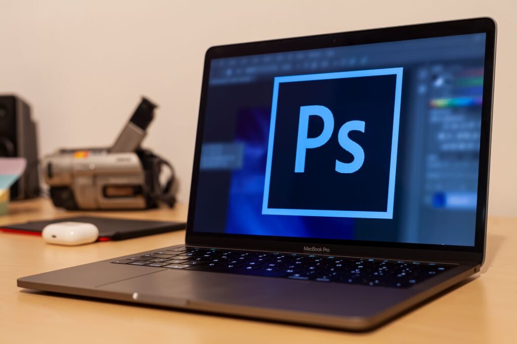 A laptop displaying Photoshop on the screen, placed on a desk with digital and office equipment.